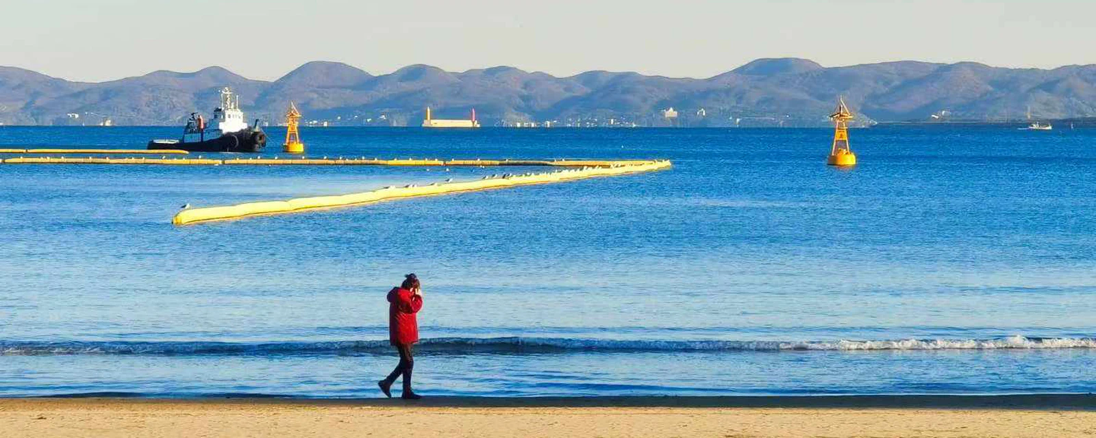 A quiet shoreline with a walker, tugboat, and distant hills